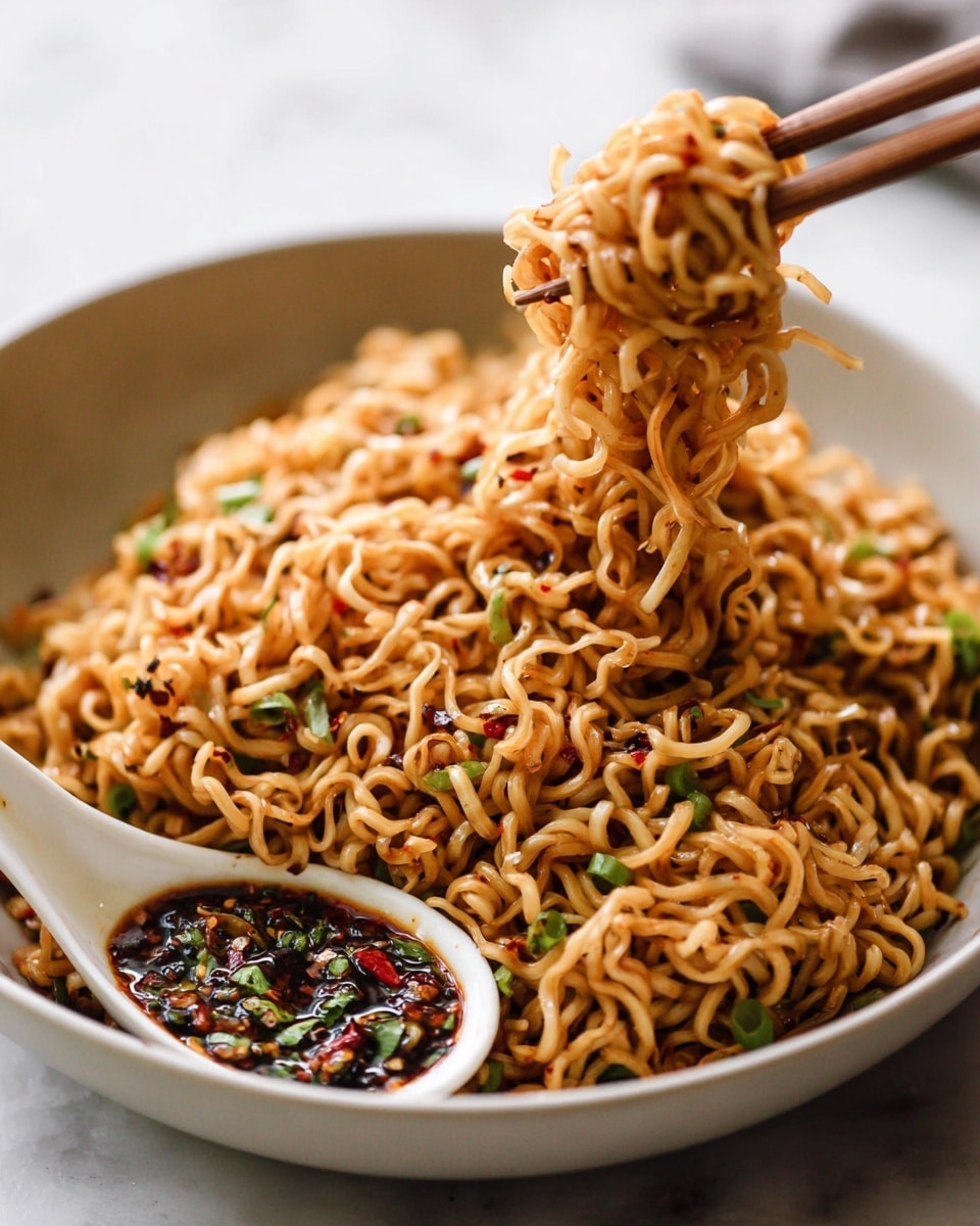 A close-up of a large pile of cooked noodles in a white bowl, with a glossy, slightly oily texture, mixed with small bits of green onions and red chili flakes scattered throughout. The noodles are tangled and twisted, showing a golden-brown color with darker spots from the seasoning. A pair of wooden chopsticks lifts a thick clump of noodles from the bowl, showing the dense, soft texture. Next to the noodles, a white ceramic spoon rests inside the bowl, holding some dark, oily chili sauce with green herbs. The whole scene is set on a white marbled surface. photo taken with an iphone --ar 4:5 --v 7