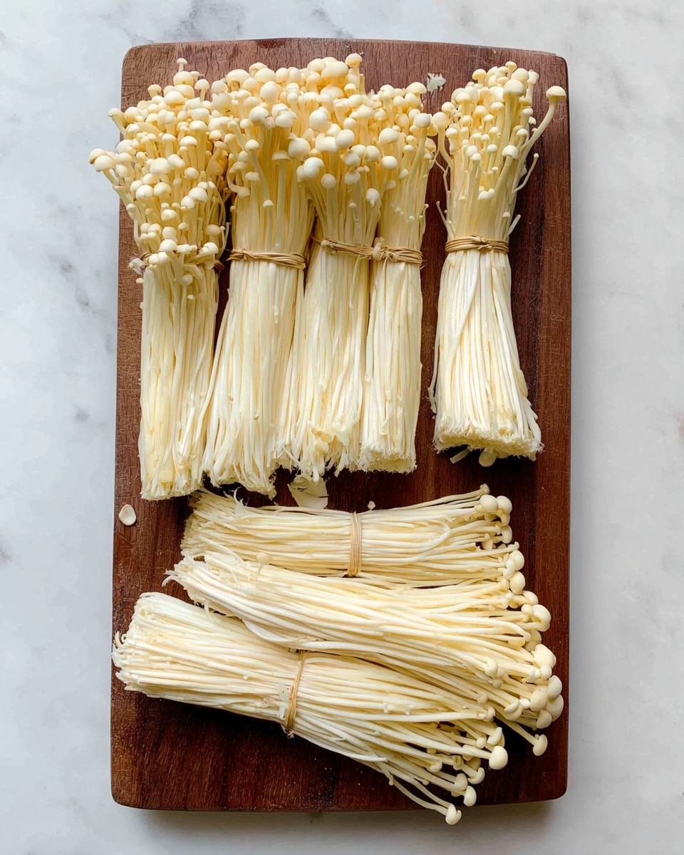 The image shows bundles of pale yellow enoki mushrooms arranged in two rows on a brown chopping board, with most clusters tied together and one smaller loose cluster on the right side. The long, thin mushroom stems have small round caps, and they are neatly placed parallel to each other. The chopping board rests on a white marbled surface, creating a clean and simple background. No additional objects or colors are visible in the image photo taken with an iphone --ar 4:5 --v 7