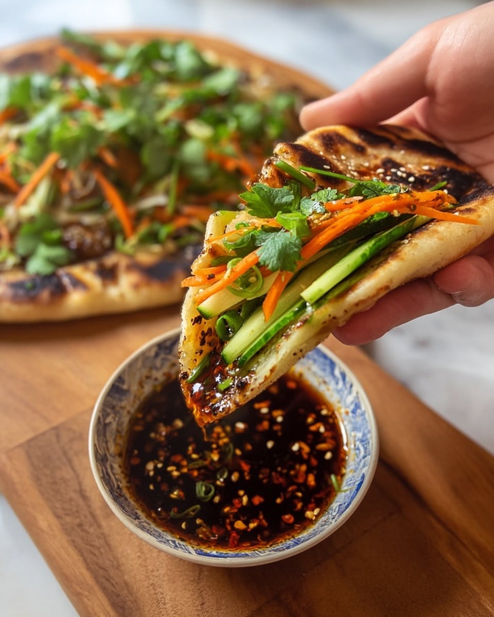 A woman's hand holds a flatbread sandwich filled with bright green cucumber strips, thin orange carrot pieces, fresh green herbs like cilantro, and thinly sliced green onions, all layered on a golden-brown, slightly charred flatbread. The sandwich is being dipped into a small bowl of dark reddish-brown chili oil with visible chili flakes and sesame seeds. In the blurred background on a wooden surface, there is another similar flatbread topped with the same fresh vegetables and herbs. The scene is set on a white marbled texture surface. Photo taken with an iphone --ar 4:5 --v 7