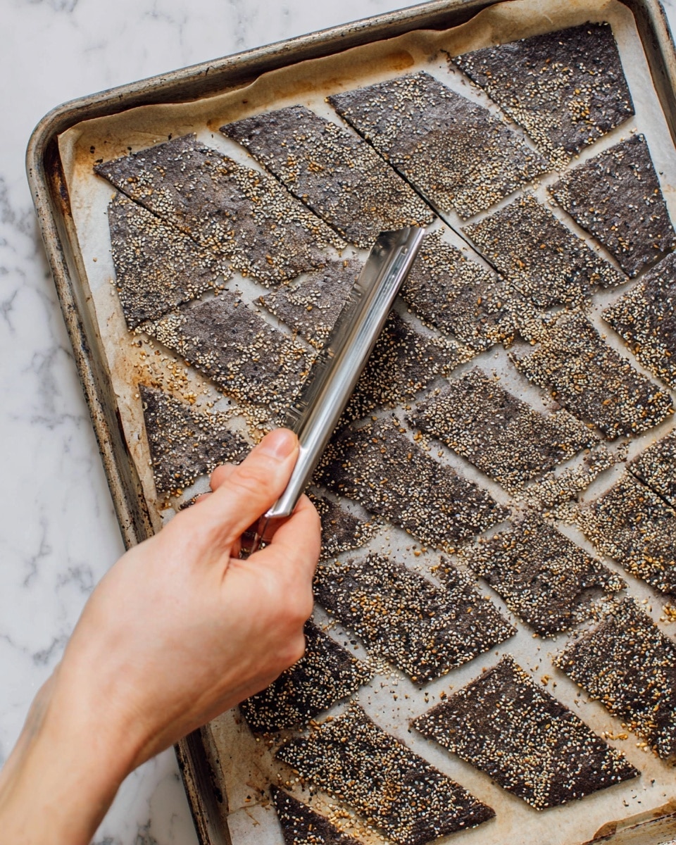 A close-up shows a thick dark gray cracker dough baked on a baking sheet lined with parchment paper, sprinkled evenly with white, black, and golden toasted sesame seeds and spices. The dough is marked with shallow diamond-shaped or triangular cuts, creating a textured pattern across the surface. A woman's hand holds a shiny metal tool pressing into the dough, emphasizing the cuts. The edges of the baking sheet are slightly stained from use, with a white marbled surface beneath it. photo taken with an iphone --ar 4:5 --v 7