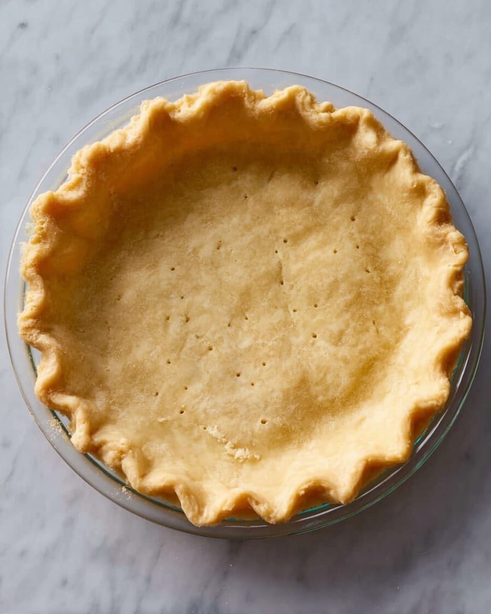 A single round pie crust with a golden-brown color fills a clear glass pie dish. The crust has a slightly crimped edge all around, creating a soft wavy pattern. Small fork holes are evenly sprinkled across the flat bottom of the crust. The pie dish sits on a white marbled surface. photo taken with an iphone --ar 4:5 --v 7