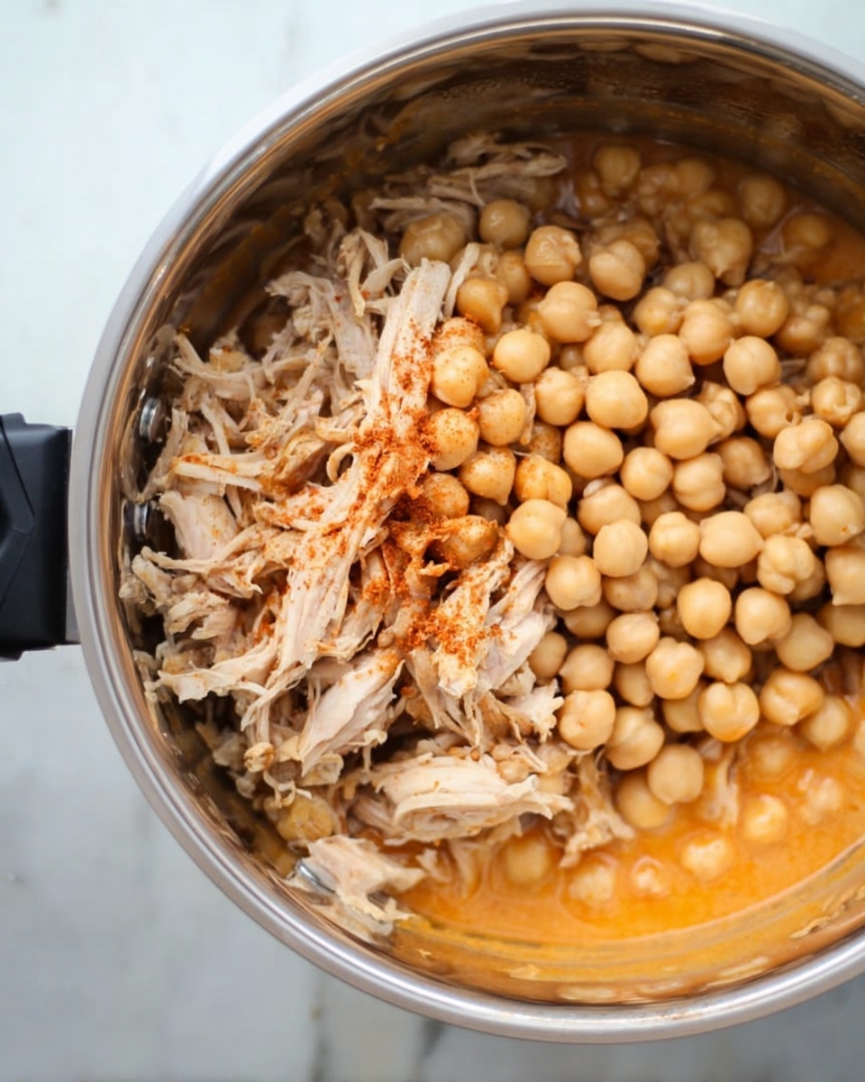 Inside a shiny metal pot, there is a layer of shredded light brown meat on one side with a slight reddish seasoning and a layer of beige chickpeas piled neatly next to it. The bottom of the pot shows a creamy light orange sauce hinting at a rich, warm flavor. The pot sits on a white marbled surface, and a black utensil handle is partially visible on the left side. photo taken with an iphone --ar 4:5 --v 7