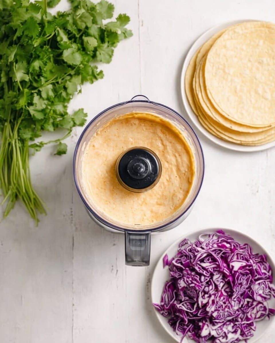 The image shows a top view of four main elements on a white marbled surface. In the center is a blender filled with a thick, light orange creamy mixture. To the left is a bunch of fresh green cilantro and below it a pile of shredded purple cabbage with white veins. On the right is a white plate holding a neat stack of pale yellow tortillas, arranged in a circle. The colors contrast well against the white marbled background. photo taken with an iphone --ar 4:5 --v 7