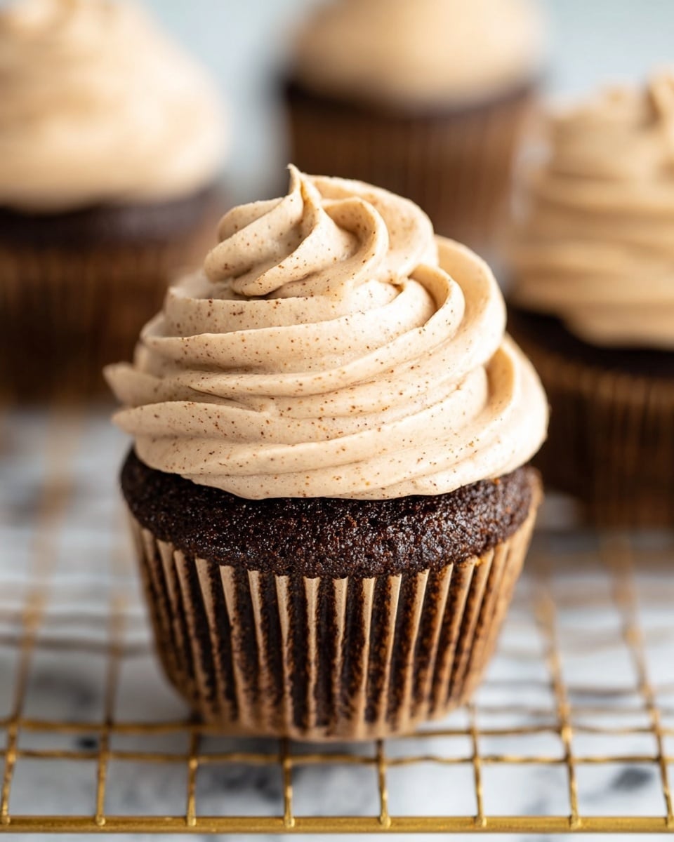 A chocolate cupcake with a deep brown, moist texture sits in a light brown paper liner. On top, there is a thick swirl of creamy light beige frosting speckled with tiny darker bits, giving it a slightly grainy look. The cupcake is placed on a gold wire cooling rack with a white marbled surface underneath. In the background, there are slightly blurred cupcakes with the same frosting and dark base. photo taken with an iphone --ar 4:5 --v 7