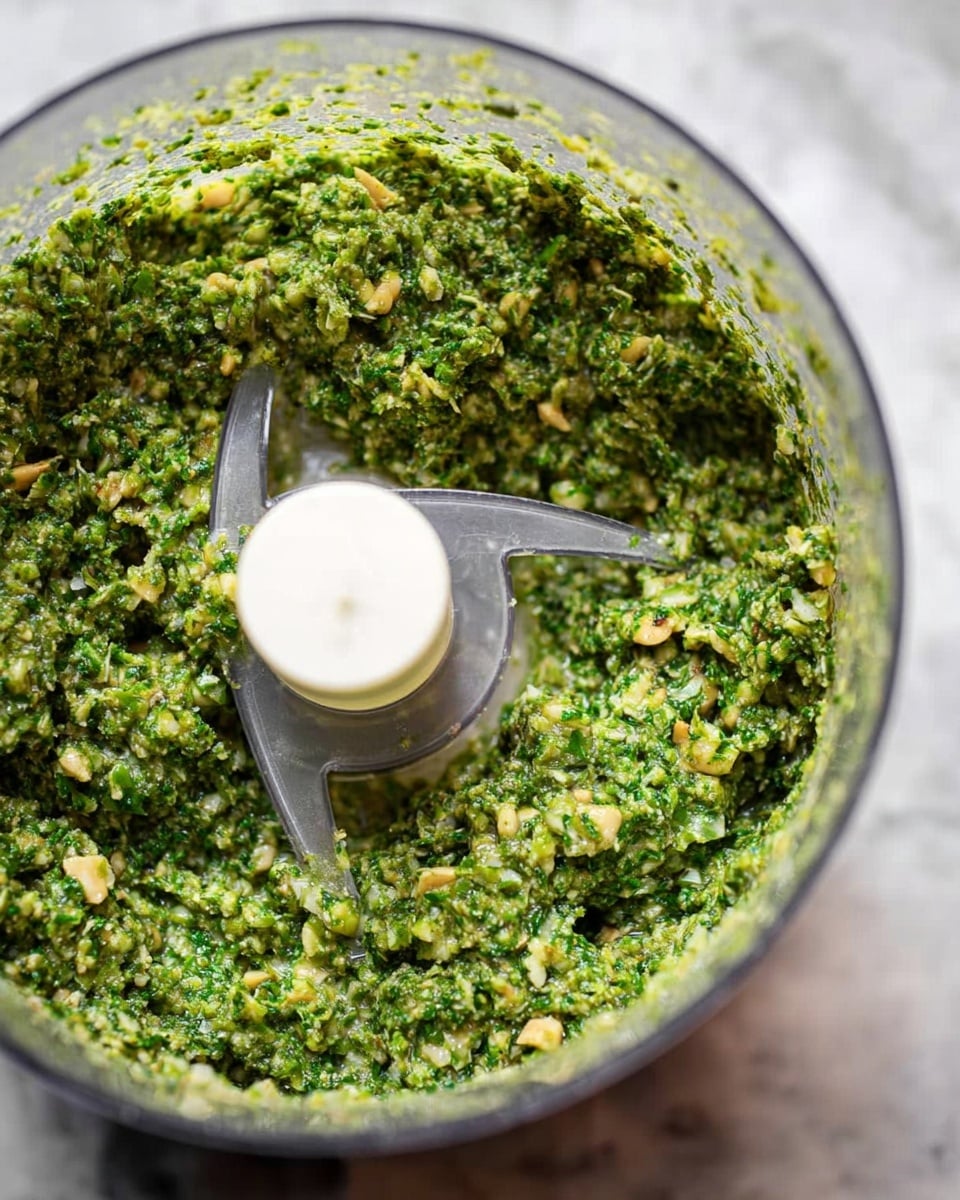 A close-up view of a clear food processor bowl filled with a thick, textured green mixture made of finely chopped herbs and nuts, showing small bits of light and dark green with some beige crumbs mixed in. The mixture covers most of the inside surface of the bowl, and the central clear plastic blade holder stands out empty and clean in the middle. The background shows a white marbled texture surface. photo taken with an iphone --ar 4:5 --v 7