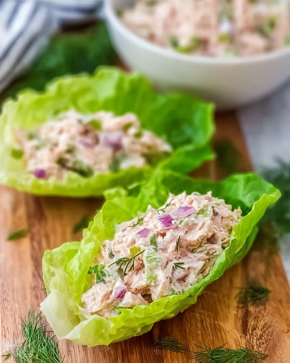 The image shows two bright green lettuce leaves used as boats, each filled with a creamy chicken salad mixture. The salad has a light pinkish-white color with small purple bits of red onion and green pieces of herbs mixed inside. The lettuce leaves are placed on a wooden board with some dill sprigs scattered around. A white bowl with more salad is blurred in the background. The scene is bright and fresh with a clear focus on the filled lettuce leaves. photo taken with an iphone --ar 4:5 --v 7