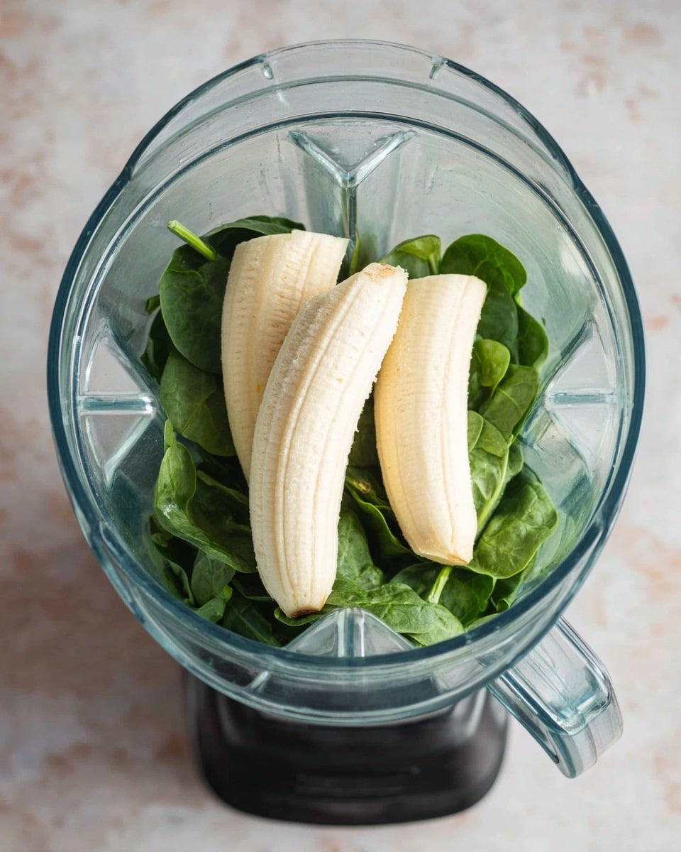 A clear blender jar with a black base filled with fresh green spinach leaves at the bottom layer, topped with three peeled bananas placed horizontally across the spinach. The background is a white marbled texture. Photo taken with an iphone --ar 4:5 --v 7