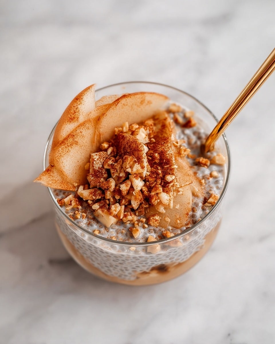 The dish is served in a transparent round glass filled with three visible layers: the bottom layer is a creamy chia seed pudding with small visible black seeds, the middle layer contains thin light brown apple slices coated with a cinnamon-like spice, and the top layer is sprinkled generously with crushed nuts and more cinnamon powder. A shiny golden spoon is partly inside the glass, resting on the right side. The glass is placed on a white marbled surface. Photo taken with an iphone --ar 4:5 --v 7