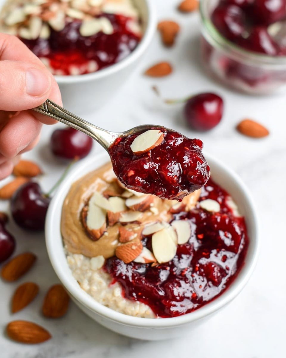 The image shows a white jar filled with three main layers: the bottom layer is light beige oatmeal, the middle layer is a thick layer of dark red chia seeds soaked in liquid, and the top layer is light beige oatmeal again. On top, there is a drizzle of white milk, a swirl of creamy brown nut butter, a dark red cherry, and some chopped light-colored almonds. In the background, there is a white bowl with oatmeal topped with red cherries and chia seeds, all on a white marbled surface with some whole and chopped almonds scattered around. Photo taken with an iphone --ar 4:5 --v 7