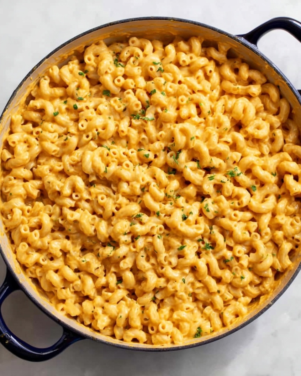 A large round pan filled with a single layer of cooked elbow macaroni pasta coated in a creamy orange cheese sauce. The pasta is evenly covered with the sauce, giving the dish a smooth and shiny texture. Small green herb bits are sprinkled on top, adding a touch of color contrast. The pan has dark handles on both sides, and it is placed on a white marbled surface. The lighting highlights the creamy texture of the cheese sauce, making the pasta look rich and comforting photo taken with an iphone --ar 4:5 --v 7
