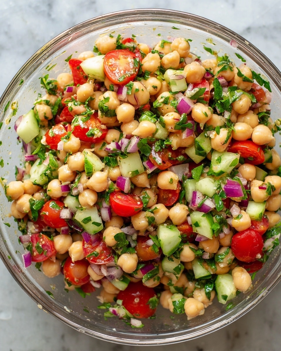 The image shows a clear bowl filled with a colorful chickpea salad. The salad has several layers and textures: light beige chickpeas that make up most of the dish, bright red cherry tomatoes cut into halves and quarters, small pieces of green cucumber, finely chopped purple-red onions scattered throughout, and fresh green parsley leaves adding a leafy texture and color contrast. All ingredients are mixed evenly, creating a vibrant and fresh look with a mix of round, smooth chickpeas and crunchy diced vegetables. The bowl sits on a white marbled surface, and the photo taken with an iphone --ar 4:5 --v 7