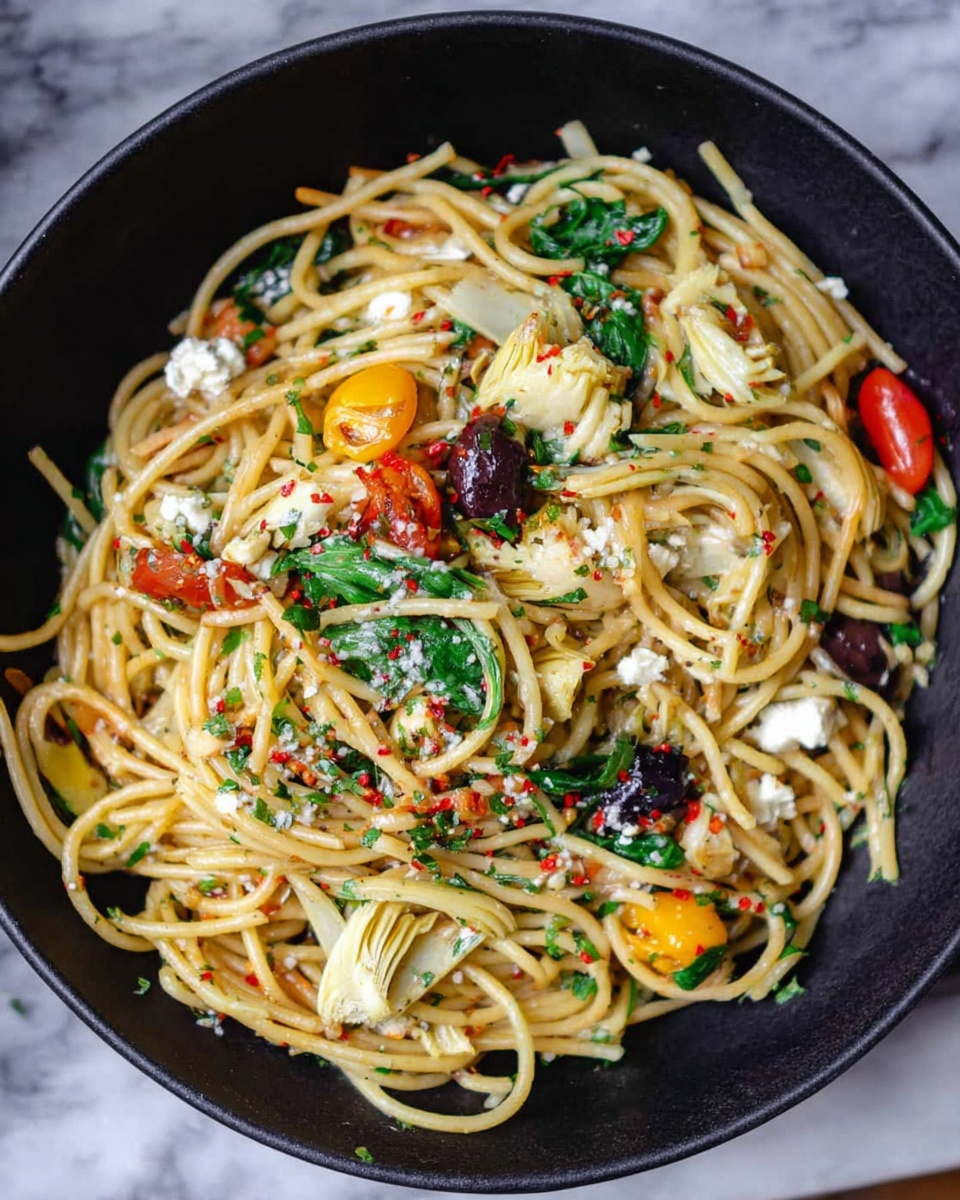 Two white plates hold servings of spaghetti pasta mixed with bright red cherry tomato halves, scattered green basil leaves, white crumbled cheese, and pale artichoke pieces. The yellow pasta strands twist and layer on the plates, topped with small red pepper flakes and bits of dark olives. A silver fork is placed on one plate with a woman's hand partially visible holding it. Next to the plates is a small white bowl filled with red pepper flakes. The scene is set on a white marbled textured surface. photo taken with an iphone --ar 4:5 --v 7