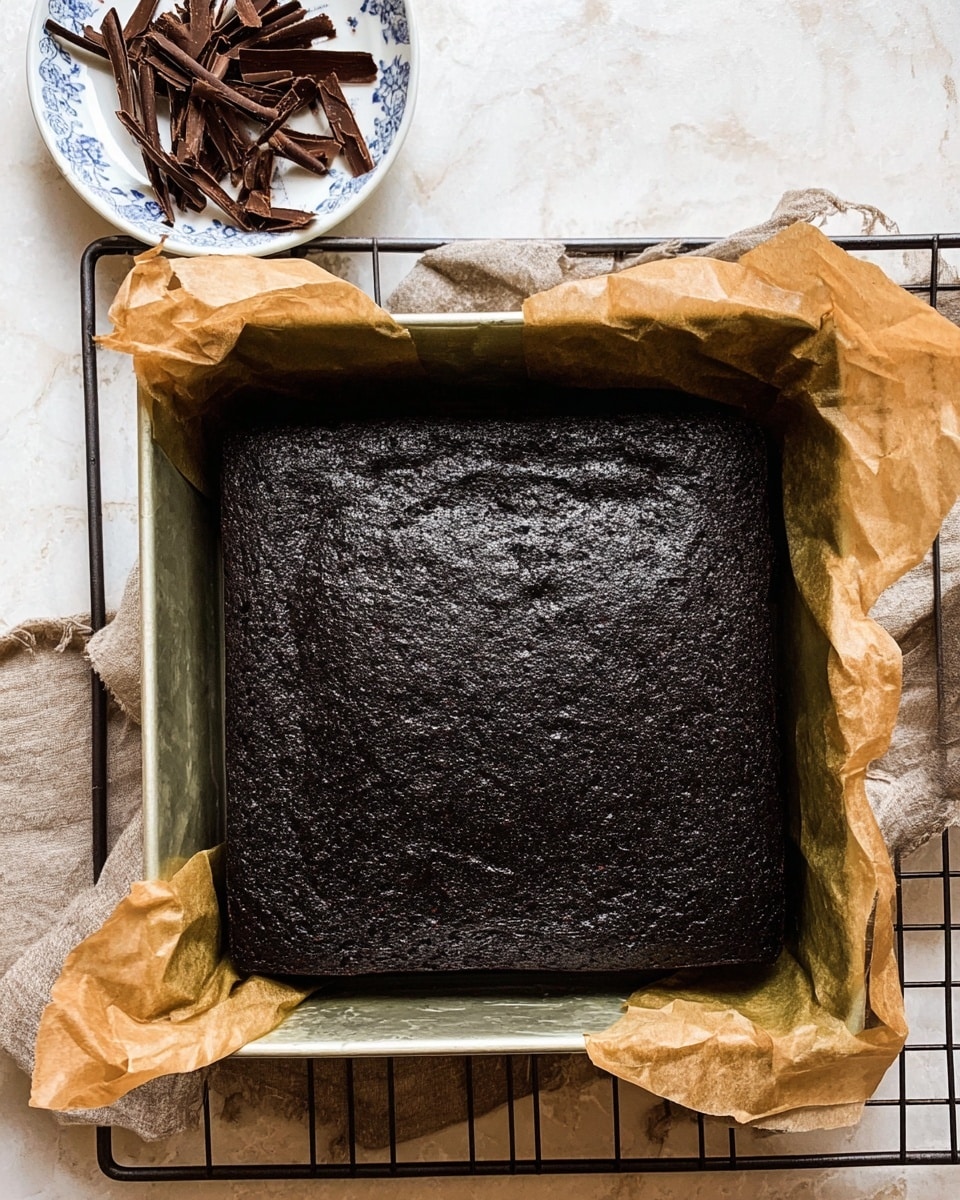 A dark black square cake with a slightly shiny and cracked surface sits inside a metal baking pan lined with crumpled brown parchment paper. In the upper left corner of the image, there is a white plate with a blue floral pattern, holding several thin, curled chocolate shavings. The baking pan is placed on a wrinkled sheet of parchment paper set on a wire cooling rack, all against a white marbled texture background. photo taken with an iphone --ar 4:5 --v 7