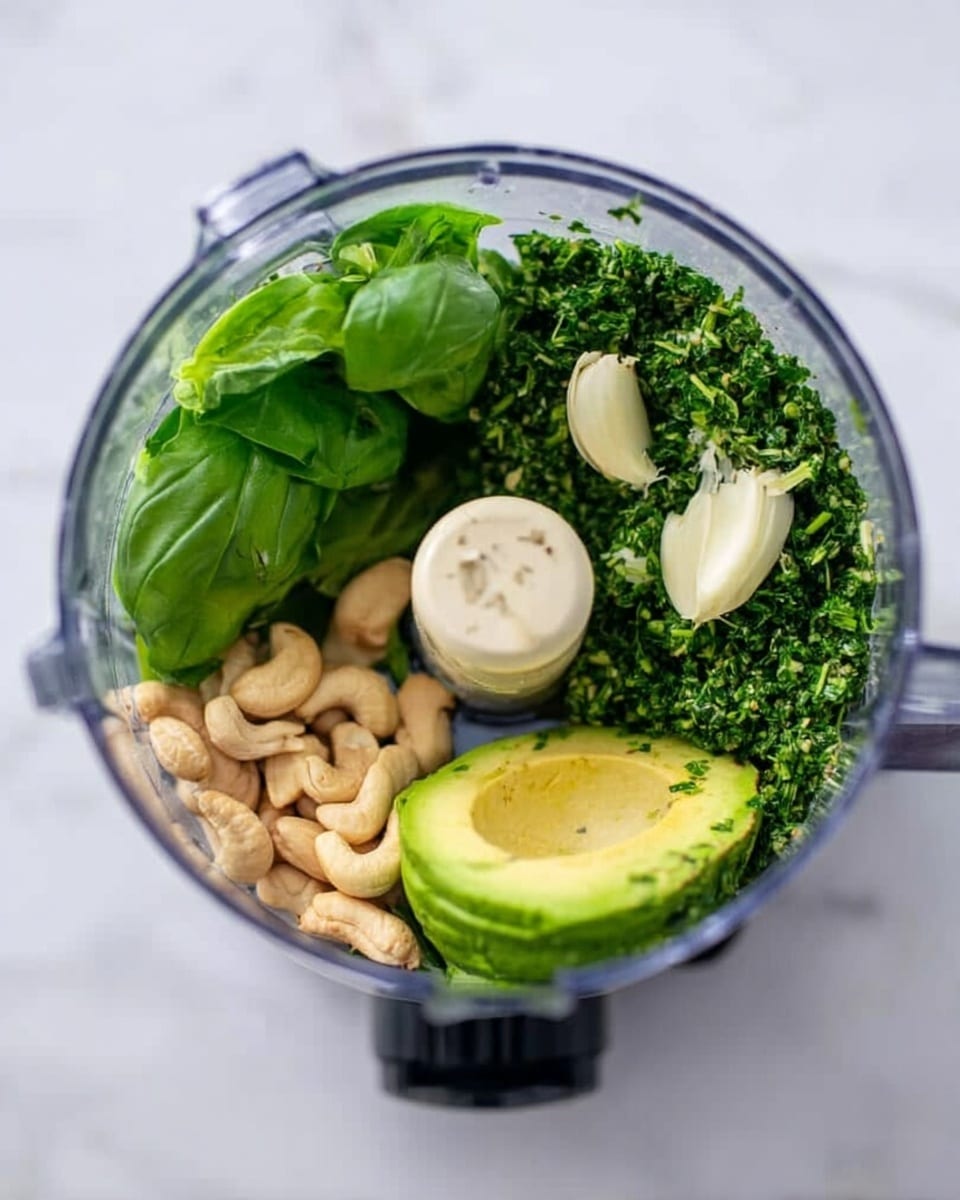 The image shows a clear food processor bowl viewed from above, containing layers of fresh ingredients. On the left side, there are several bright green basil leaves with a smooth and shiny texture. Next to them is a darker green pile of chopped herbs, possibly parsley or cilantro, with a finer texture. On the right side, two peeled garlic cloves sit on top of the herbs. Below the garlic, two halves of an avocado with light green flesh and a creamy texture are placed side by side. At the bottom center, there is a small cluster of light-colored cashew nuts. The food processor is on a white marbled surface. photo taken with an iphone --ar 4:5 --v 7