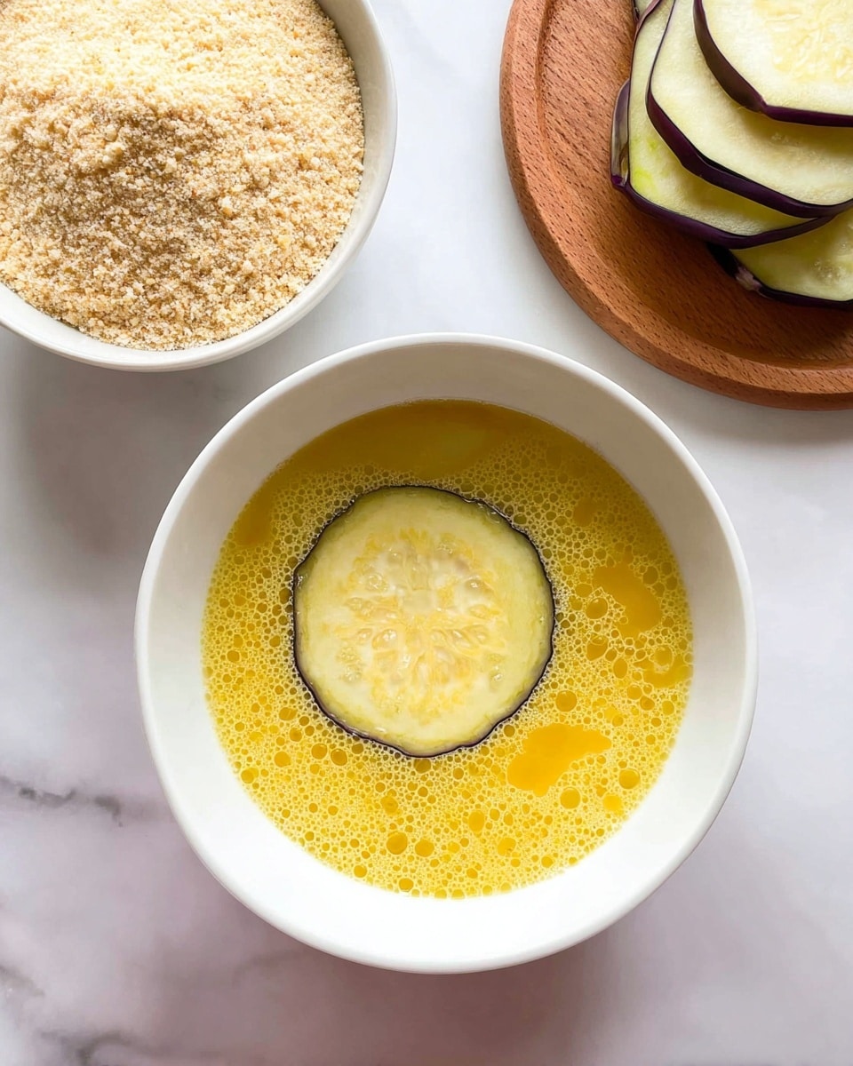 The image shows a white bowl centered on a white marbled surface, filled with yellow beaten eggs with bubbles on the surface; a round light purple eggplant slice with darker edges floats in the middle of the egg mixture. Above and slightly to the left, there is another white bowl filled with light beige breadcrumbs that have a grainy texture. To the right edge of the image, part of a wooden board is visible, holding several stacked slices of the same purple eggplant. photo taken with an iphone --ar 4:5 --v 7