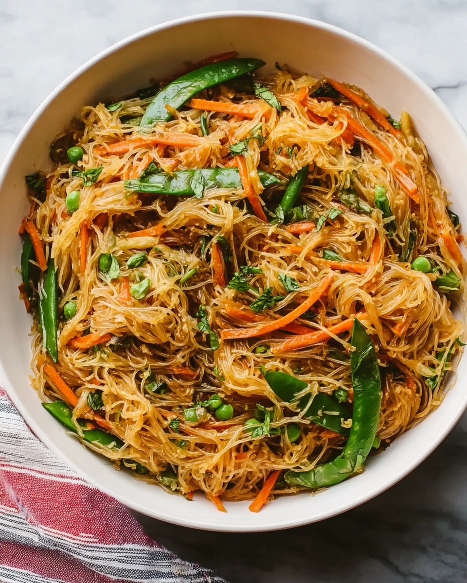A white bowl filled with thin, light brown noodles mixed with orange carrot strips and bright green leafy vegetables. The noodles and vegetables are mixed evenly, creating a colorful and textured look. A woman's hand is holding dark brown chopsticks lifting some noodles and a bit of the vegetables above the bowl. The bowl sits on a white marbled surface, with a soft blue blurred background behind. photo taken with an iphone --ar 4:5 --v 7
