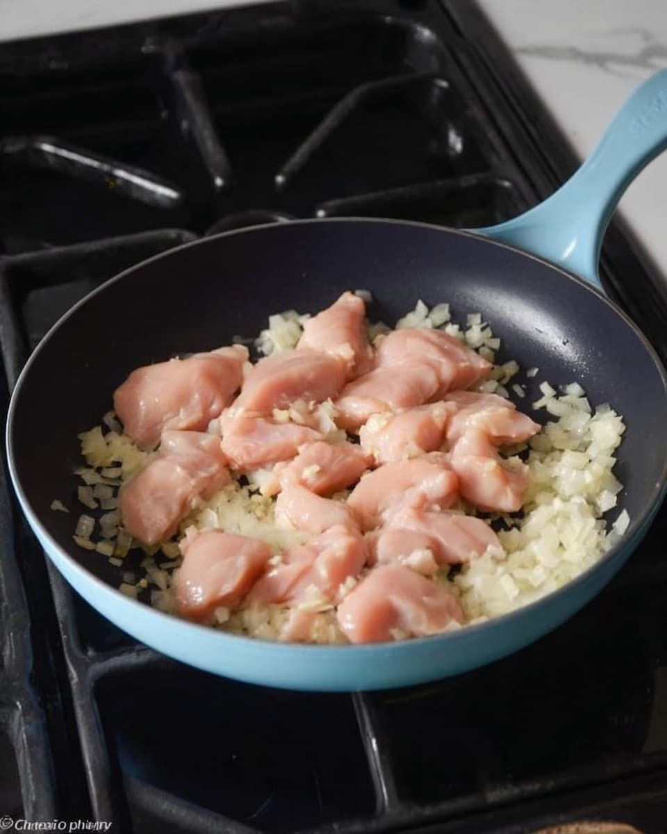 In a light blue skillet on a stove with black grates, there are small pieces of raw light pink chicken placed on top of finely chopped white onions that are slightly translucent, spread evenly on the bottom of the pan. The chicken pieces have smooth, shiny surfaces and are scattered across the pan, with some garlic pieces mixed with the onions. The black inside of the skillet contrasts with the colors of the chicken and onions. The background shows parts of the stove in dark tones, all set on a white marbled surface. photo taken with an iphone --ar 4:5 --v 7