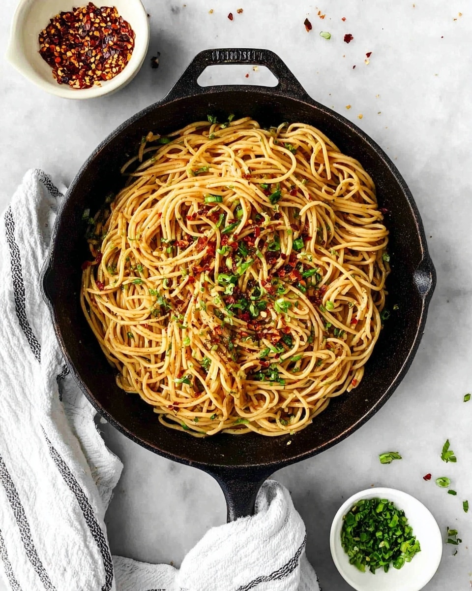 A black cast iron skillet filled with long, light brown noodles topped with small pieces of red chili flakes and bright green chopped herbs, all mixed together evenly. The skillet rests on a white marbled surface, and a white and gray striped cloth is wrapped around the skillet handle. Next to the skillet, there are two small white bowls, one containing red chili flakes and the other with chopped green herbs. Some green herbs are scattered on the white marbled surface around the skillet. photo taken with an iphone --ar 4:5 --v 7