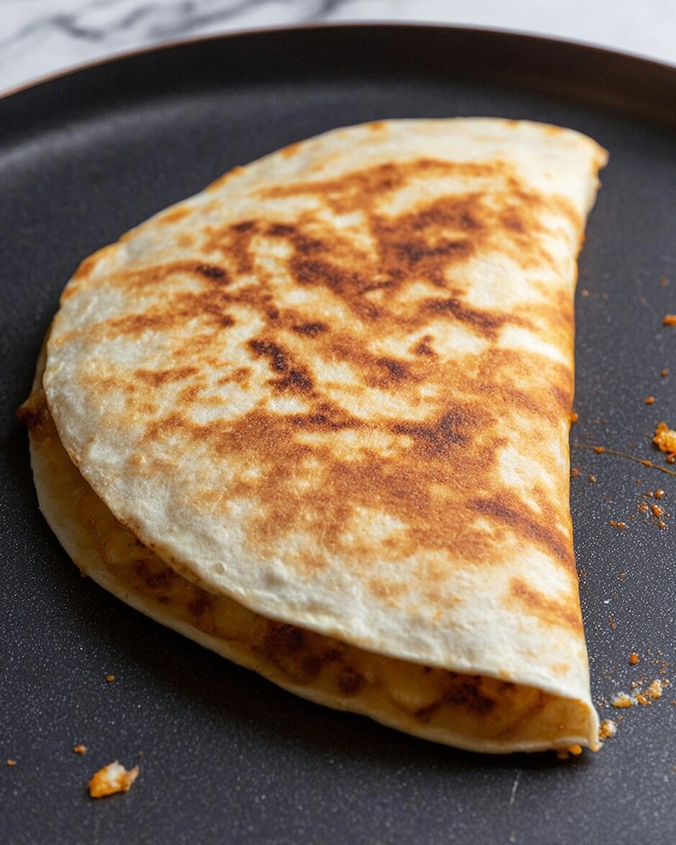 A folded, lightly toasted quesadilla with a golden-brown surface showing even but varied browning across its top layer. The tortilla has a soft, slightly puffed texture with visible edges sealed tightly, hiding the filling inside. Small bits of orange-colored filling poke out slightly near the edge. The quesadilla is placed flat on a dark, non-stick pan with a few scattered crumbs around it. The background has a white marbled texture. photo taken with an iphone --ar 4:5 --v 7