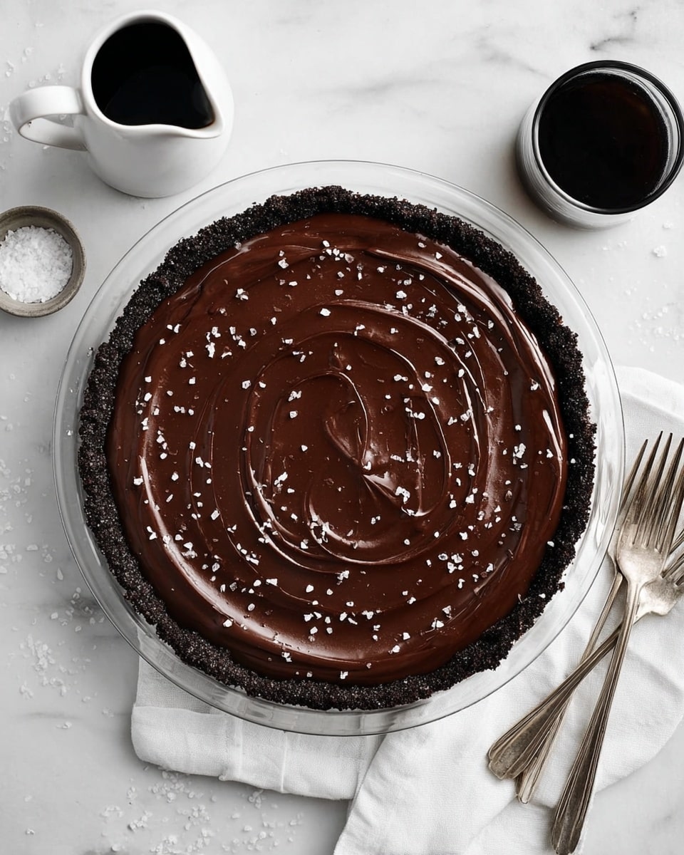 A round pie with a dark crumbly crust forms the outer layer, holding a smooth, shiny chocolate filling that has gentle swirling patterns on top. The top is lightly sprinkled with small white flakes, adding texture. The pie sits on a transparent cooling rack, which rests on a white marbled surface. Nearby are two silver forks on the right and a white pitcher filled with dark liquid on the upper left, alongside a small white container with coarse salt. The scene is brightly lit and clean, showing simple, elegant details. photo taken with an iphone --ar 4:5 --v 7