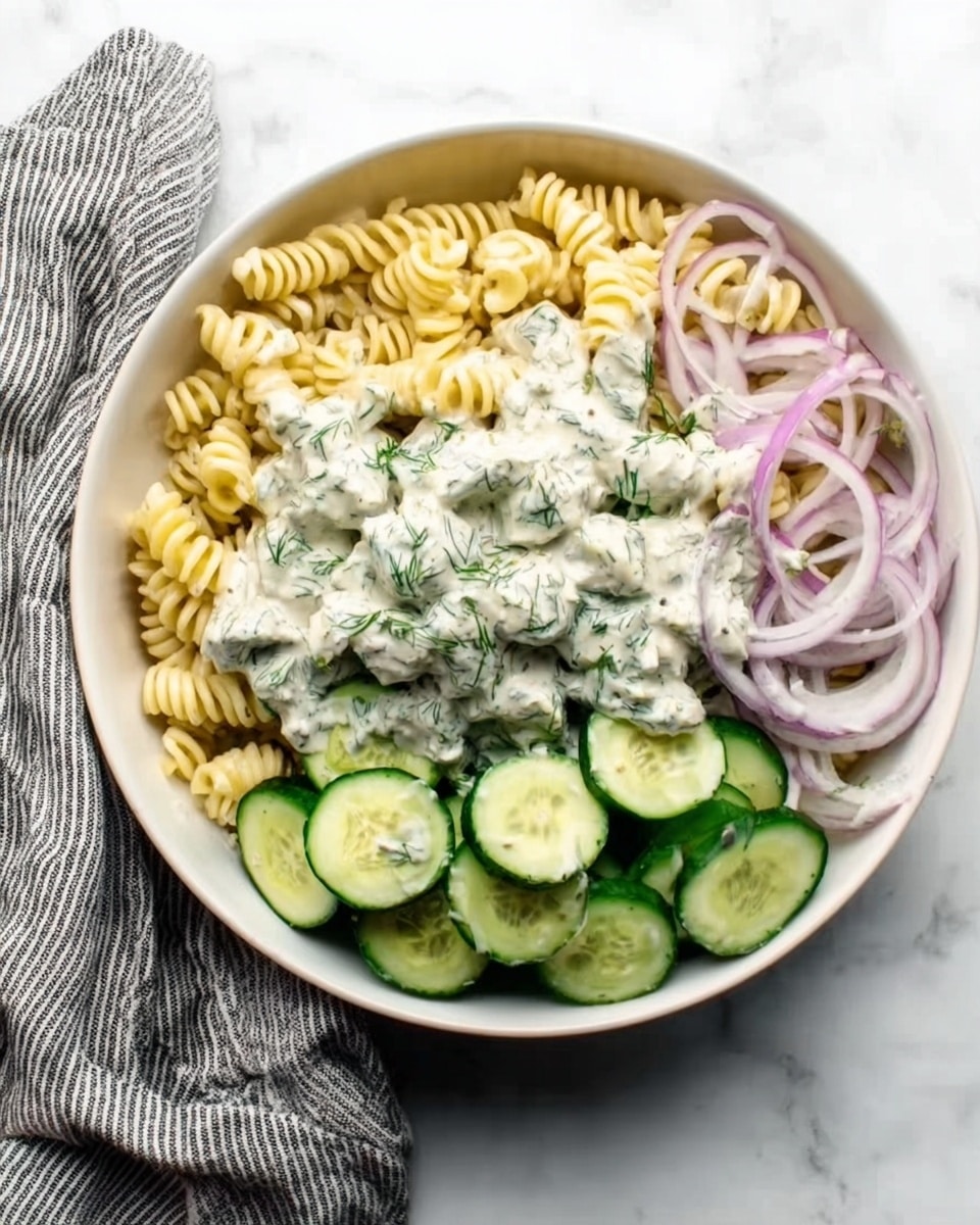 A white bowl sits on a white marbled surface filled with three main sections. One section has curly cooked pasta in a pale yellow color. Another section is filled with thin cucumber slices showing a green outer skin and light green inside. The last section has thin rings of light purple onions. On top of all the sections is a creamy white sauce with green herb bits spread evenly, adding texture and freshness. A woman's hand with a striped gray and white cloth is near the bowl. Photo taken with an iphone --ar 4:5 --v 7