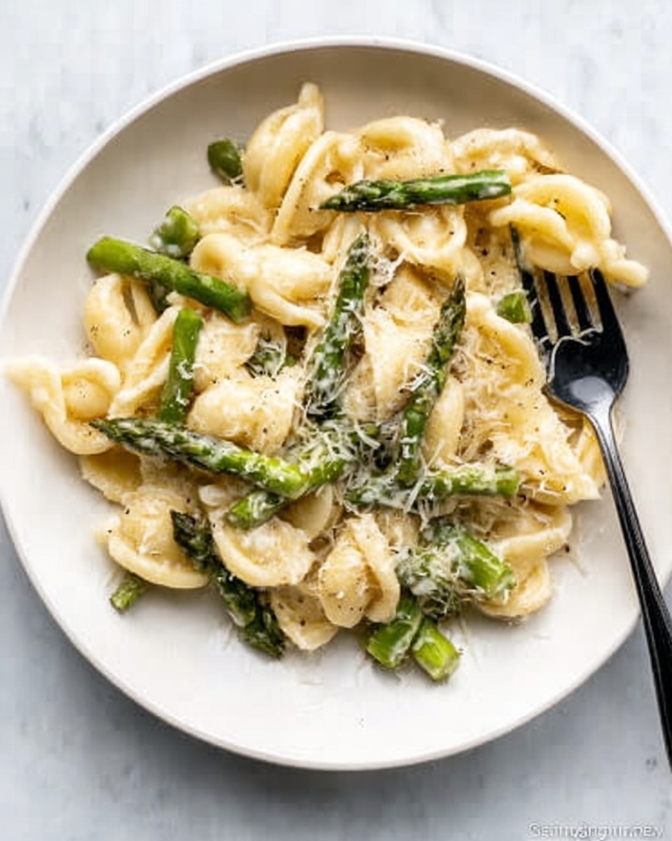 A white plate holds a serving of creamy pasta with thick, curved noodles that are pale yellow. Mixed throughout the pasta are bright green pieces of asparagus, cut into small segments. The dish is topped with a light sprinkling of finely grated white cheese. A black fork rests on the right side of the plate with some pasta and asparagus on its prongs. The plate sits on a white marbled surface. Photo taken with an iphone --ar 4:5 --v 7