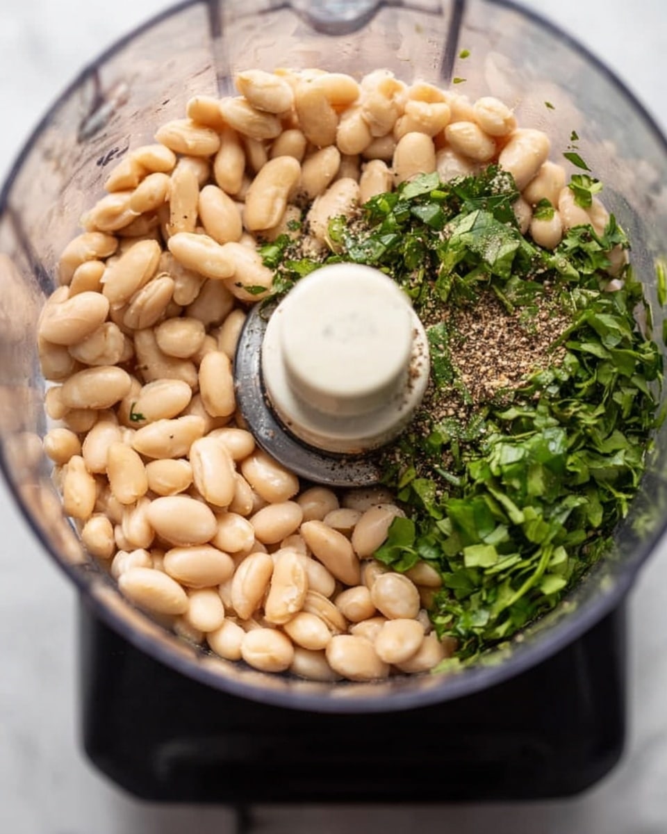A close-up view inside a clear food processor bowl showing a single smooth, creamy mixture with pale yellow and light green tones, filling about half of the bowl. The shiny metal blade is centered and partially covered by the thick mixture, which has a soft, slightly textured surface. The bowl sits on a white marbled surface with small gray and black specks scattered around. Photo taken with an iphone --ar 4:5 --v 7