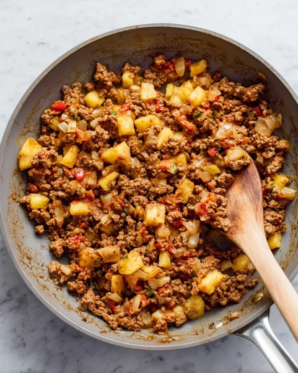 A large gray pan filled with cooked ground meat mixed with small pieces of yellow pineapple, white onions, and diced tomatoes. The meat is brown and crumbly, while the pineapple adds bright yellow chunks, and the onions appear soft and translucent. A wooden spoon is in the center of the pan, stirring the mixture. The pan sits on a white marbled surface, with natural light creating soft shadows. photo taken with an iphone --ar 4:5 --v 7