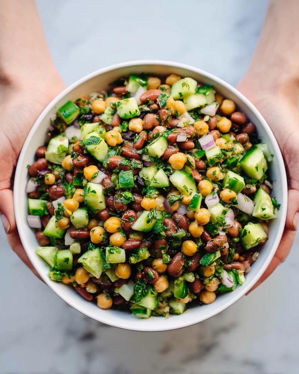 A white bowl filled with a colorful salad held by two woman's hands. The salad has three main layers: a mix of light brown beans, dark brown beans, and round yellow chickpeas forming a textured base, scattered evenly throughout. Bright green cucumber pieces are cut into small chunks and spread all over, adding freshness. Finely chopped green herbs and small white onion pieces are mixed in, enhancing the salad with more color and texture. The bowl is held above a white marbled surface. photo taken with an iphone --ar 4:5 --v 7