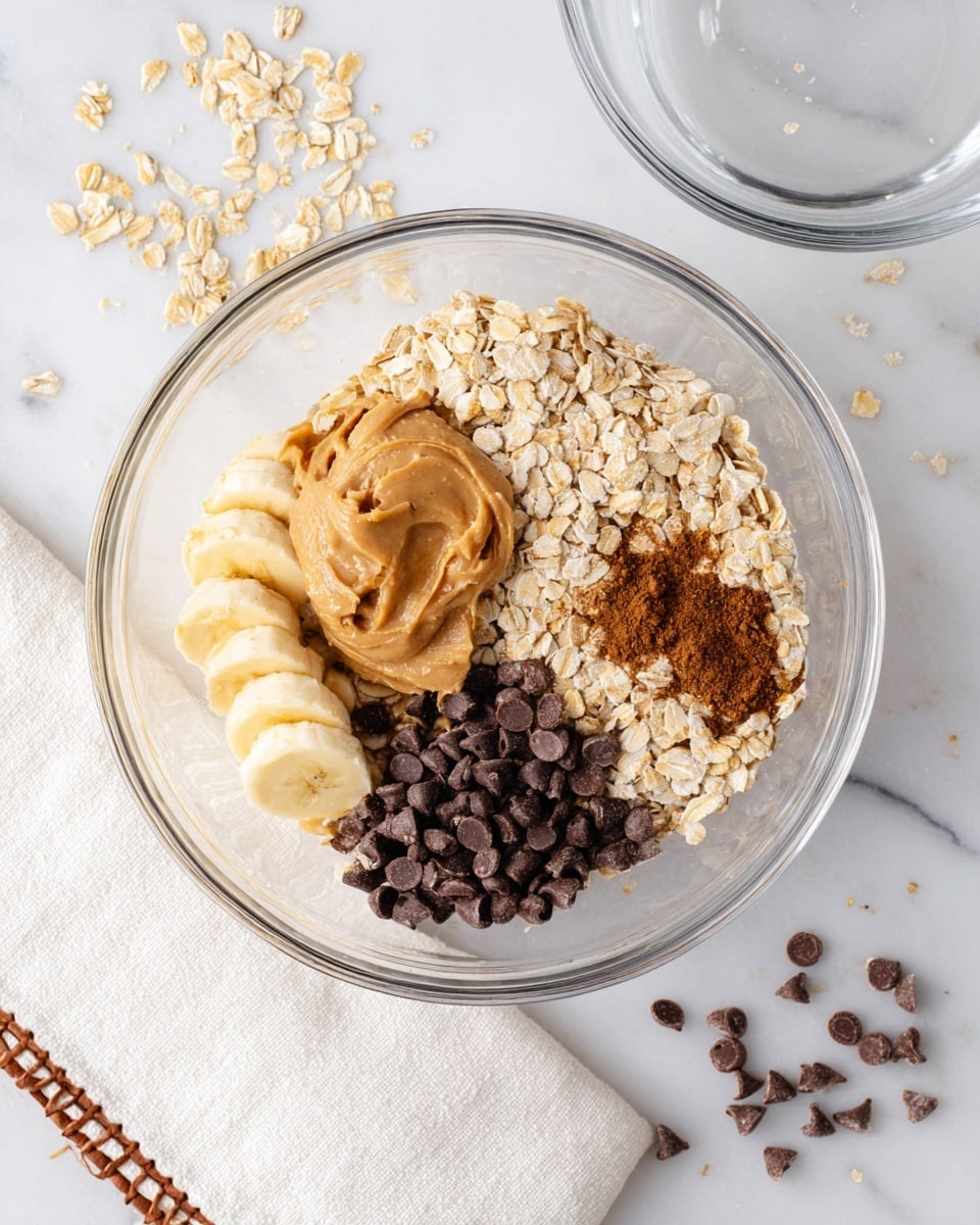 A clear glass bowl sits on a white marbled surface filled with five separate layers of ingredients: mashed pale yellow bananas at the bottom left, creamy light brown peanut butter on top of the bananas, light beige rolled oats covering the right half of the bowl, a small pile of dark brown cinnamon powder near the center on the oats, and a heap of dark brown chocolate chips at the bottom right of the oats. Tiny scattered chocolate chips and oat flakes are also on the white marbled surface nearby. Another empty clear glass bowl is above the main bowl. A white cloth with brown stitching is visible at the bottom left corner. Photo taken with an iphone --ar 4:5 --v 7