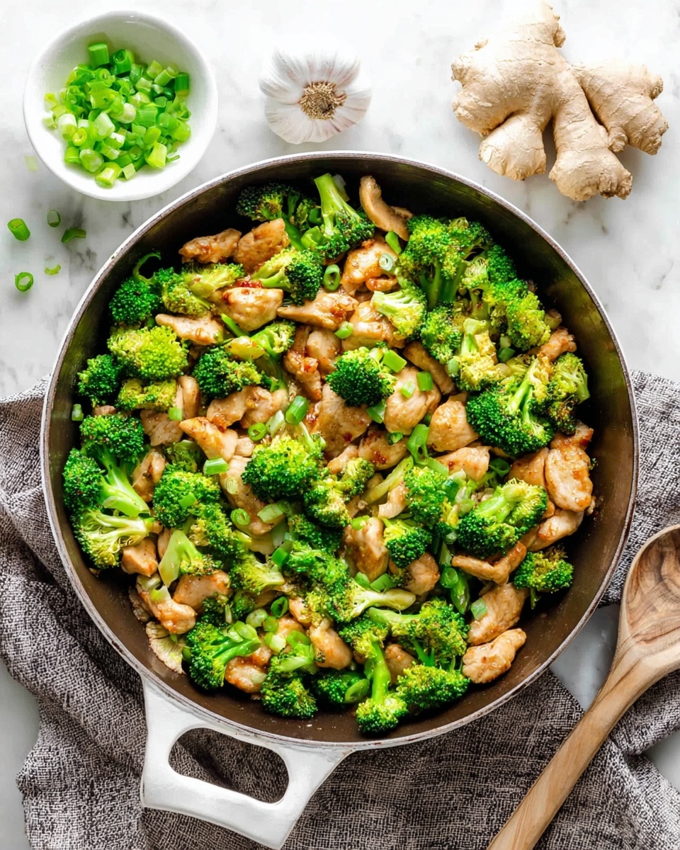 A white pan filled with a stir-fry of bright green broccoli florets and light brown cooked chicken pieces, mixed evenly throughout. The broccoli is fresh and vibrant with a slightly bumpy texture, while the chicken pieces are tender with a smooth surface and golden-brown edges. Small pieces of chopped green onions are scattered on top adding a fresh green color contrast. The pan rests on a white marbled surface, nearby a small white bowl filled with chopped green onions, a piece of ginger root, and a garlic bulb. A grey textured cloth is placed to the right side of the pan. photo taken with an iphone --ar 4:5 --v 7
