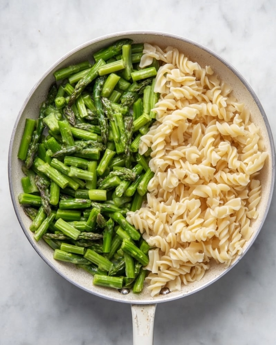 A white pan on a white marbled surface holds two distinct layers side by side: on the left, bright green asparagus pieces cut into small chunks showing a fresh, slightly glossy texture; on the right, pale beige cooked pasta with soft, twisted shapes, filling half of the pan. The colors contrast cleanly, with the green asparagus having a slight shine and the pasta appearing smooth and tender. photo taken with an iphone --ar 4:5 --v 7
