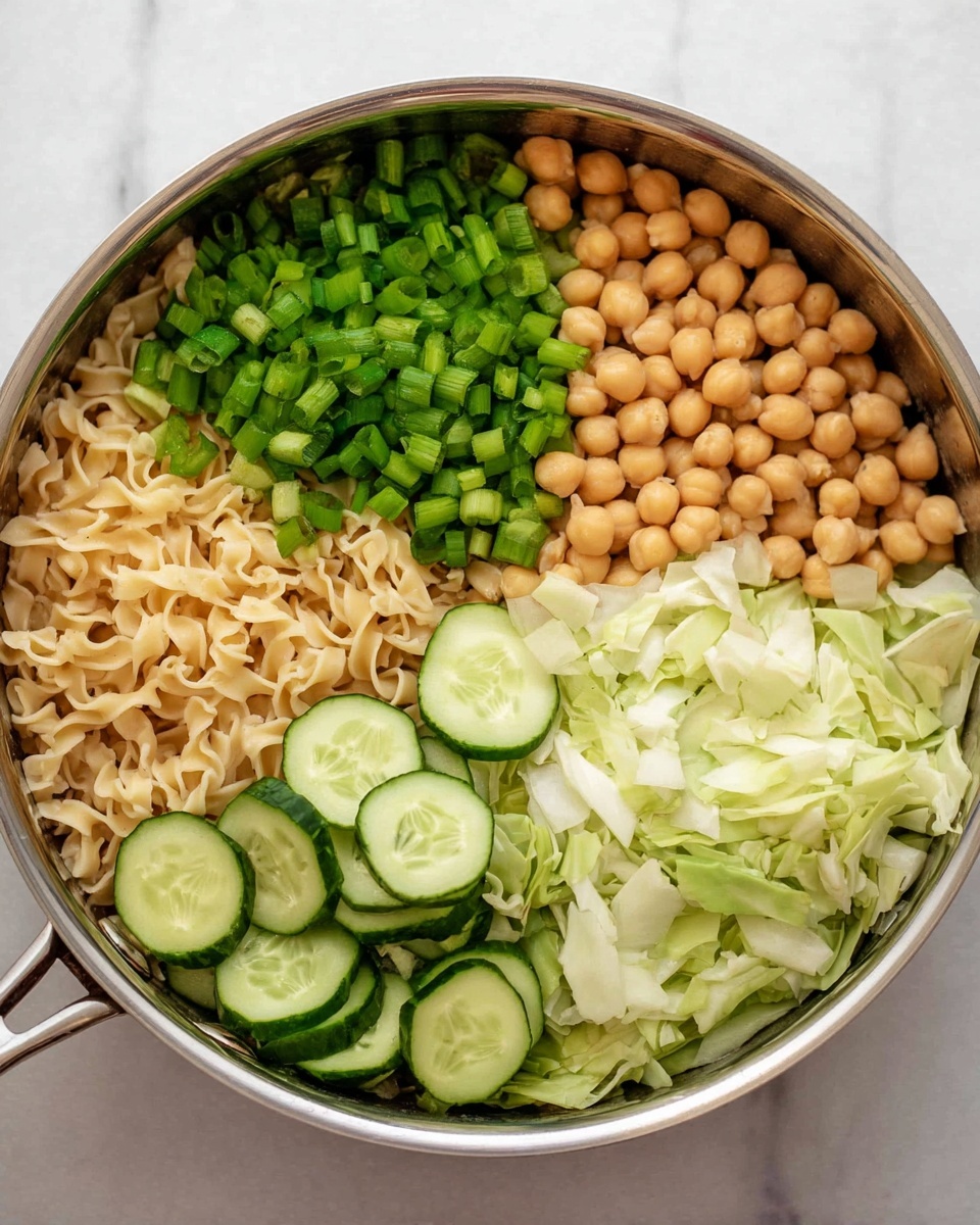 The image shows a pan filled with four main layers of ingredients arranged separately on top of each other. At the base, there is a layer of cooked curly noodles with a light beige color and soft texture. The top left section is covered with bright green chopped scallions, adding a fresh and crisp look. To the right of the scallions, there is a pile of round, light tan chickpeas with a smooth surface. Next to the chickpeas, on the right side, there is a portion of chopped white cabbage with a slightly pale green tint and crunchy texture. The bottom part of the pan is filled with thin, round slices of cucumber showing both dark green skin and light green interior. The pan sits on a white marbled surface. Photo taken with an iphone --ar 4:5 --v 7