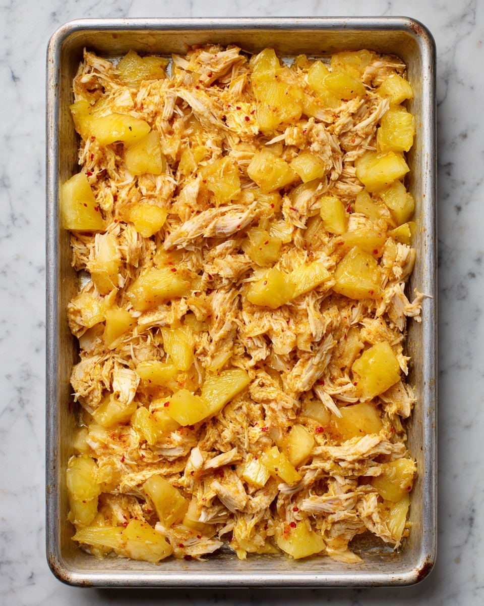 In a large metal baking tray, there is a single layer of shredded light beige chicken pieces mixed evenly with golden yellow chunks of pineapple, both spread out to cover the entire tray. The tray shows some baking marks and the food looks moist with tiny bits of red seasoning scattered throughout. The background surface is a white marbled texture. Photo taken with an iphone --ar 4:5 --v 7