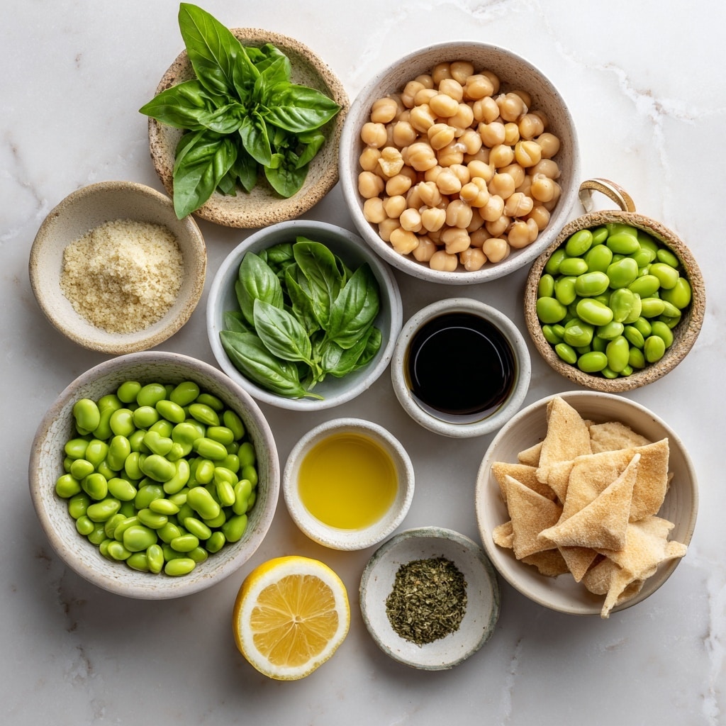 The image shows a clear food processor bowl with four main layers inside, set on a white marbled surface with a blue and white striped cloth on the right side. Starting from the top right, there is a layer of round light brown chickpeas. On the top left is a layer of bright green edamame beans. Below the edamame are three white garlic cloves. The bottom right of the bowl contains a bunch of fresh green basil leaves, some with dark veins. The center has a round white plastic part of the food processor. Photo taken with an iphone --ar 4:5 --v 7