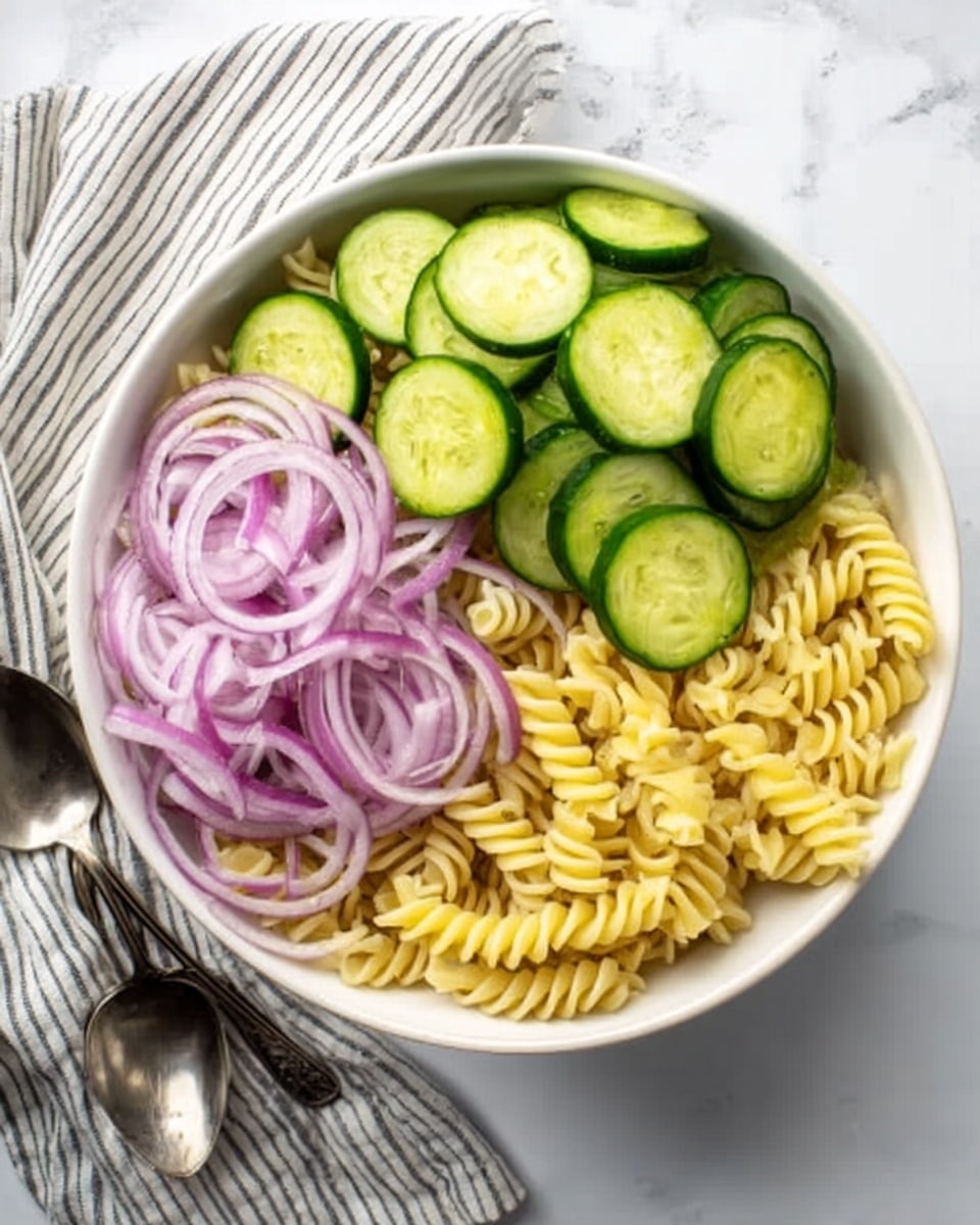 A white bowl sits on a white marbled surface, holding three main layers of food. On the right side, there is a pile of yellow spiral pasta with a smooth texture. On the left side, a layer of thinly sliced purple onions with rings stacked neatly. Above the onions, there are thinly sliced bright green cucumbers, overlapping and covering part of the pasta. A striped cloth napkin and two spoons are placed to the left side of the bowl. Photo taken with an iphone --ar 4:5 --v 7