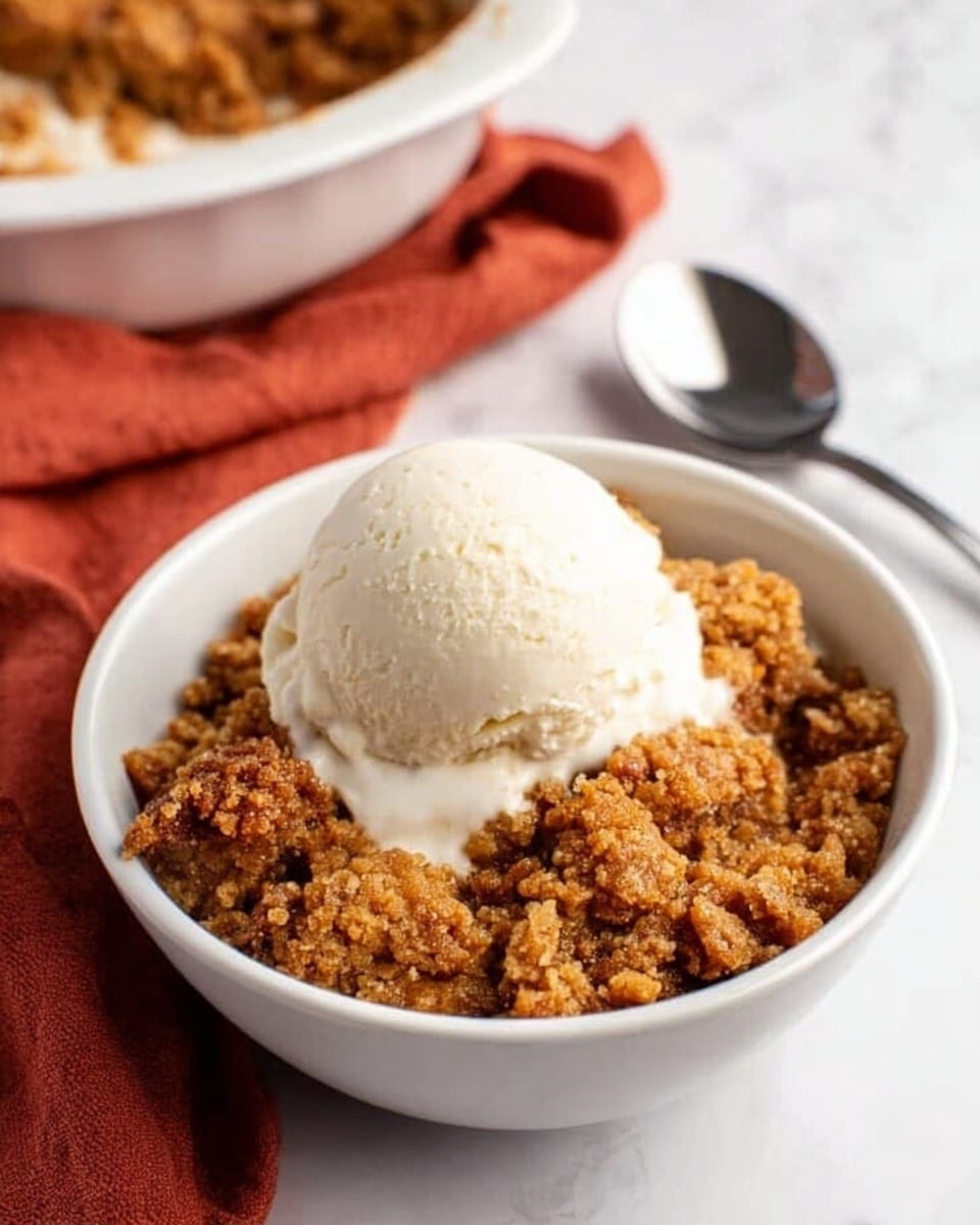 A white bowl filled with a warm, crumbly brown dessert that has a rough texture, topped with a smooth, round scoop of creamy white ice cream melting slightly on the edges. The bowl is placed on a white marbled surface with a small shiny silver spoon resting beside it. In the background, there is a partially visible white dish with more of the brown dessert and a rust-colored cloth underneath it. Photo taken with an iphone --ar 4:5 --v 7