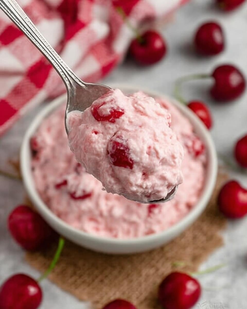 A close-up view of a silver spoon holding a large scoop of light pink creamy mixture with small red cherry pieces visible inside. The background shows a bowl filled with the same pink mixture on a textured brown surface with scattered fresh cherries. A white and red checkered cloth is partially visible on the left side, and beneath everything is a smooth white marbled surface. Photo taken with an iphone --ar 4:5 --v 7