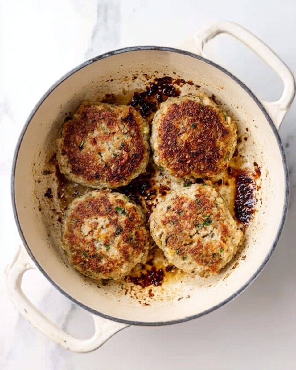 The image shows a white pot with four round, browned patties inside, each with a slightly crispy and textured surface. The patties have uneven golden brown spots and small green bits, likely herbs, throughout them. The bottom of the pot has dark browned areas from cooking, contrasting with the lighter, creamier white sides of the pot. The pot is placed on a white marbled surface and has two handles on the sides. There is a subtle shadow under the pot, giving depth to the image photo taken with an iphone --ar 4:5 --v 7
