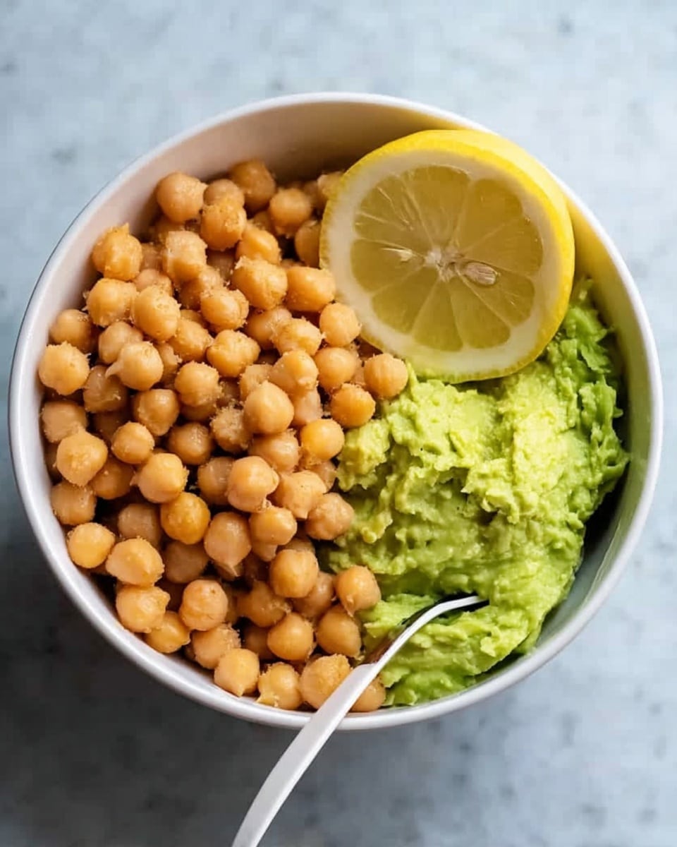 A white bowl filled with three main layers: the top left side has round, light brown chickpeas piled high, the bottom right side contains mashed avocado with a smooth green texture, and a half lemon with bright yellow skin and visible pulp rests on top next to the avocado. A white spoon is placed inside the bowl on the right side. The bowl sits on a white marbled surface. photo taken with an iphone --ar 4:5 --v 7