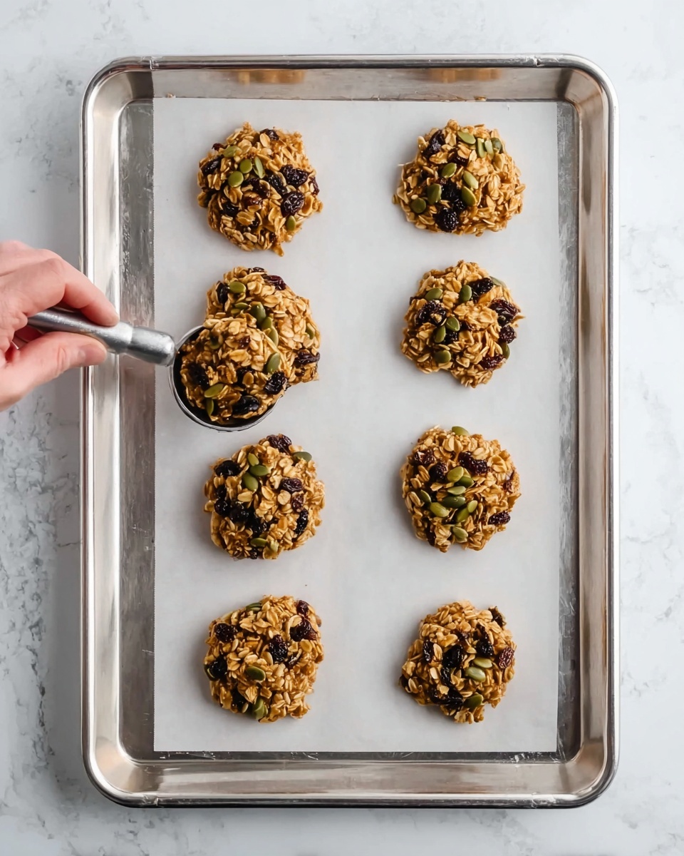 The image shows a metal baking tray lined with white parchment paper placed on a white marbled surface. On the tray, there are eight round clusters of oats mixed with dark raisins and green pumpkin seeds, spaced in two neat columns of four. Each cluster is roughly the same size, with a bumpy texture from the oats and seeds. A woman's hand using a metal cookie scoop is adding the last cluster to the bottom left corner of the tray. The overall look is natural and homey, with a clear focus on the rustic oat and seed mixture. photo taken with an iphone --ar 4:5 --v 7
