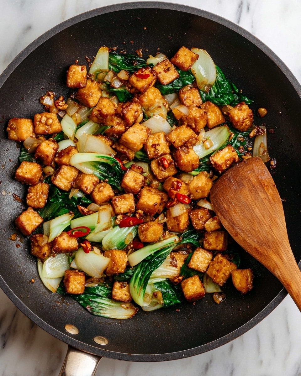 A black frying pan filled with small golden-brown crispy tofu cubes mixed with light green bok choy leaves showing some dark green near the edges, scattered with thin slices of pale onion and small slices of red chili. A wooden spatula is resting on the right side inside the pan. The pan sits on a white marbled surface. photo taken with an iphone --ar 4:5 --v 7