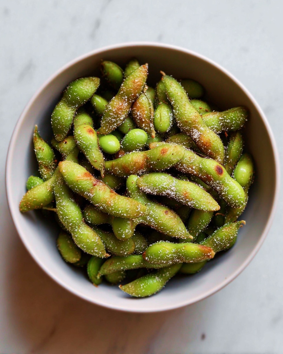 A white bowl filled with cooked green edamame pods that have a slight shine and are coated with small red and brown spices. The edamame pods look soft and slightly wrinkled, piled on top of each other, with some pods showing curved shapes and different sizes. The bowl sits on a white marbled surface near a window with bright natural light shining on the edamame, creating soft shadows. photo taken with an iphone --ar 4:5 --v 7