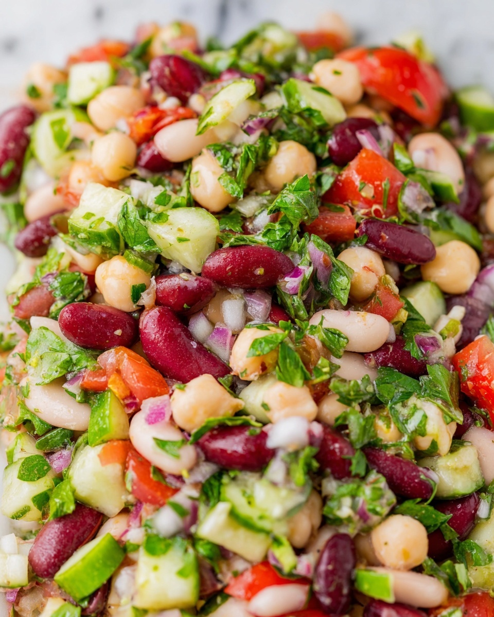 The image shows a close-up of a mixed bean salad with many colorful ingredients. There are three main layers: bright red kidney beans, creamy white beans, and small round chickpeas. Mixed in are pieces of chopped green cucumber, bright red tomato, and small bits of purple onion. Fresh green herbs like parsley or cilantro are spread evenly throughout, adding a fresh texture. The salad looks fresh and healthy with a mix of soft beans and crunchy vegetables on a white marbled surface. photo taken with an iphone --ar 4:5 --v 7