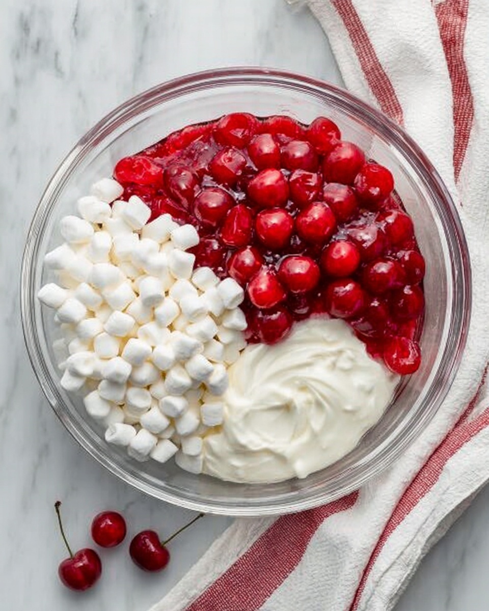 A clear glass bowl is placed on a white marbled surface, filled with three distinct layers. One section shows bright red cherries with vibrant juice, the next is full of small, soft white marshmallows stacked neatly, and the last section has smooth, creamy white yogurt with a thick texture. The bowl is slightly covered on the right side by a white cloth with red stripes, and a few loose cherries are scattered around on the marble. Photo taken with an iphone --ar 4:5 --v 7
