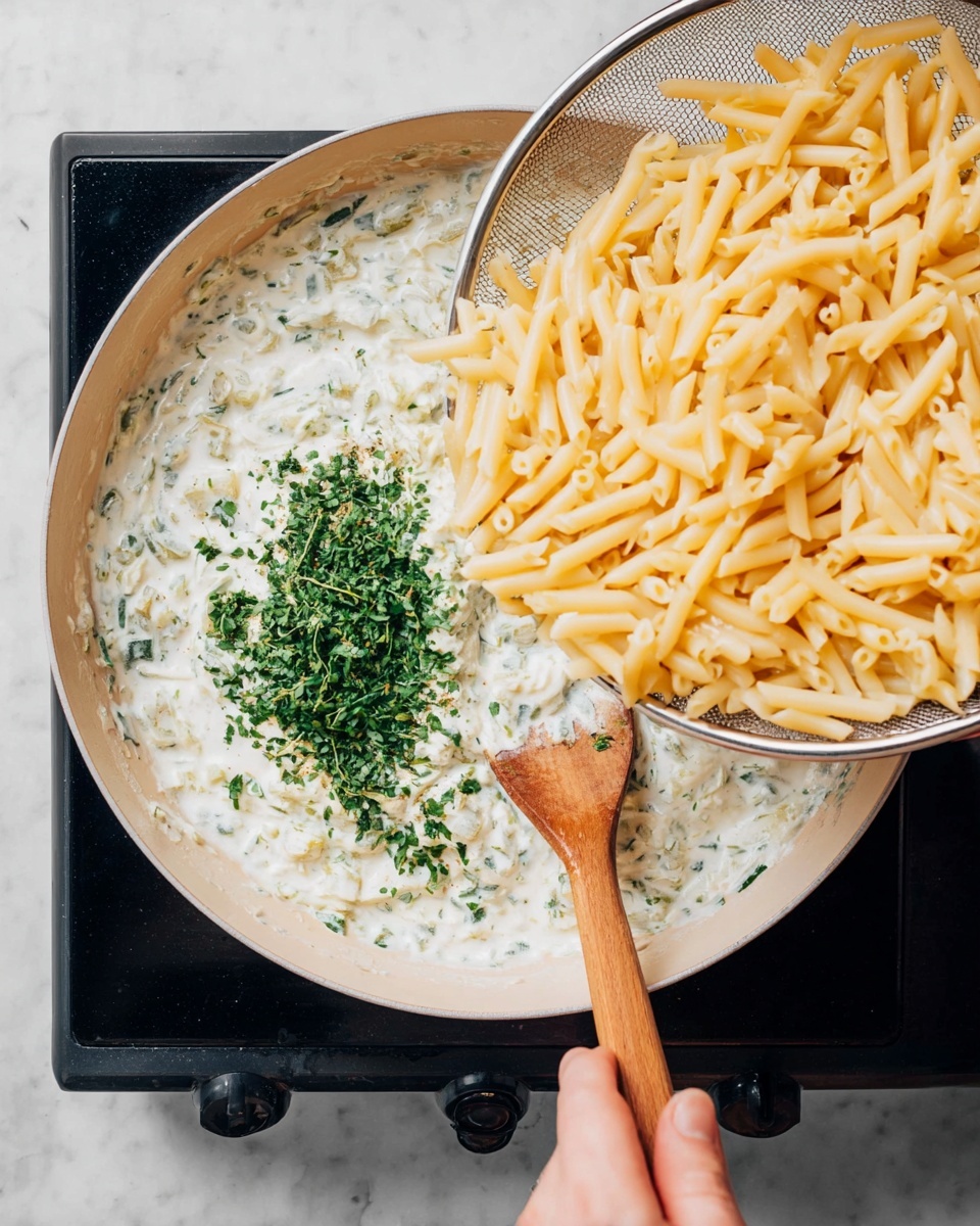 A creamy white sauce mixed with finely chopped green herbs fills one side of a large, light-colored pan on a black stovetop, while a pile of pale yellow, cooked macaroni pasta is being added from a metal strainer held by a woman's hand on the right side. A wooden spoon lays on the sauce side of the pan, resting diagonally. The background is a white marbled texture. photo taken with an iphone --ar 4:5 --v 7