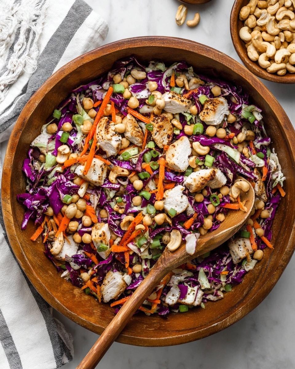 A large wooden bowl filled with a colorful salad shows several layers and textures: the base is made of chopped purple cabbage with dark purple and white tones, topped with shredded orange carrot strips, light beige chickpeas, and small pieces of grilled white chicken with brown grill marks. Scattered green slices of spring onions and whole cashews with a creamy beige color add texture. A wooden spoon is placed inside the bowl for serving. This bowl sits on a white marbled surface with a white and black striped cloth partially visible in the top left corner and a small wooden bowl with extra cashews in the top right corner. photo taken with an iphone --ar 4:5 --v 7